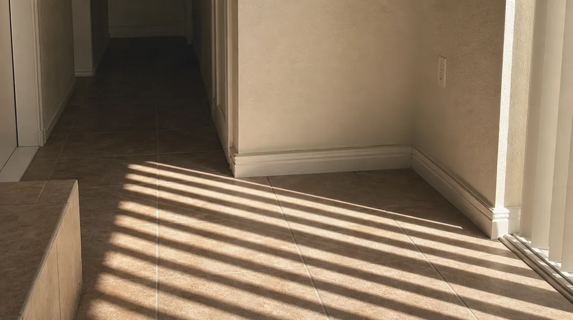 A tiled hallway corner in hard afternoon light, the floor running unbroken to a doorway with no visible source of water